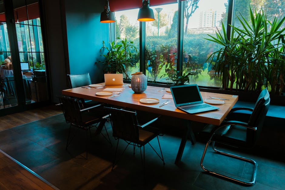 A modern dining room setup with laptops, plates, and lush plants by large windows.